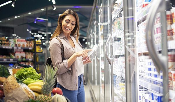 Woman with shopping list standing by the fridge in supermarket and checking cart.